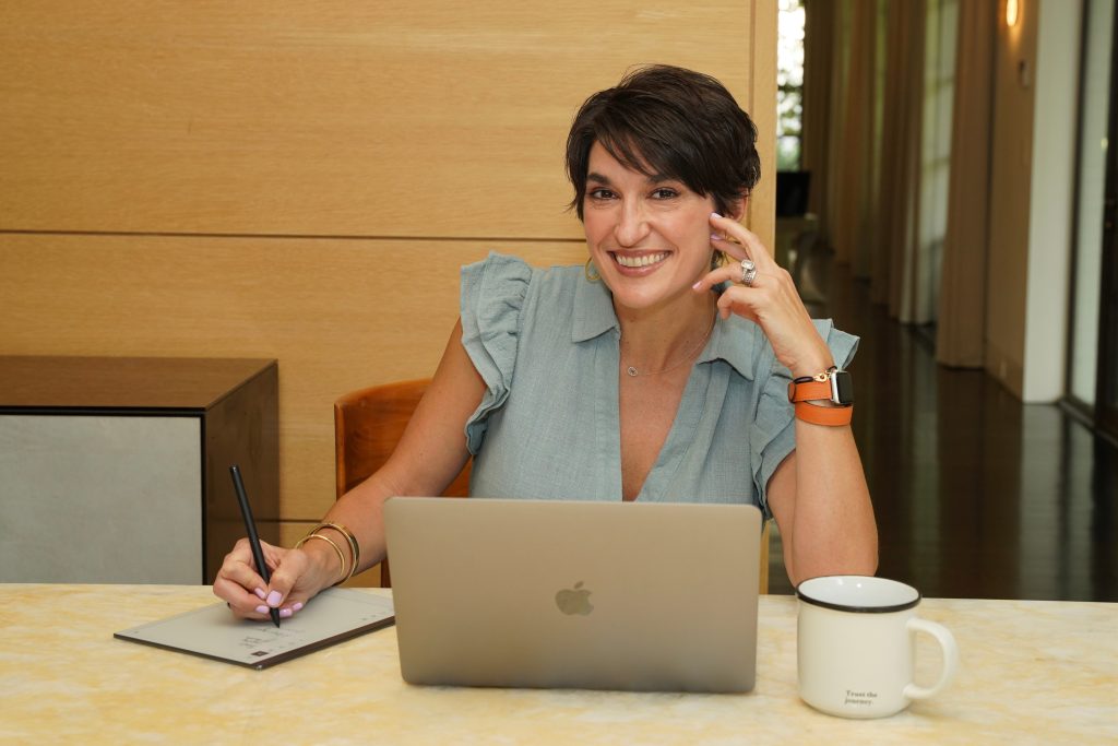 Woman smiling while taking notes beside a laptop and coffee mug at a desk.
