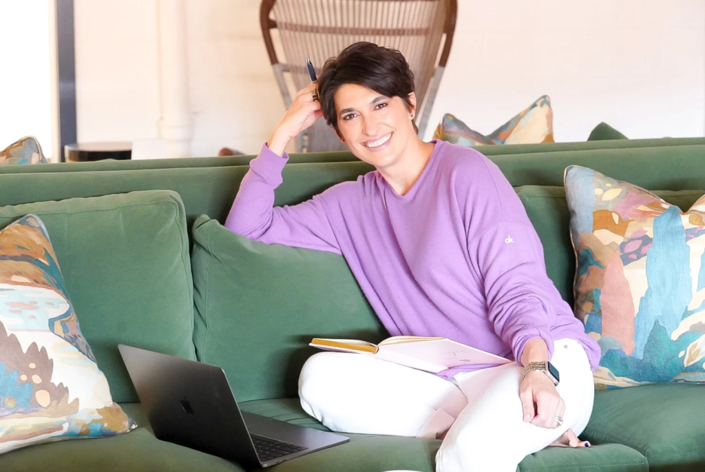 Woman smiling at camera sitting on green sofa next to a laptop with an open book in lap