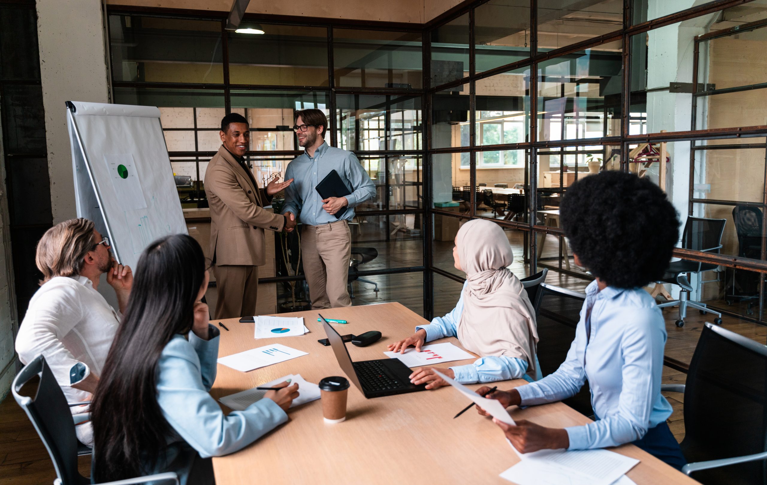 Two people shake hands in front of a table of other business professionals.