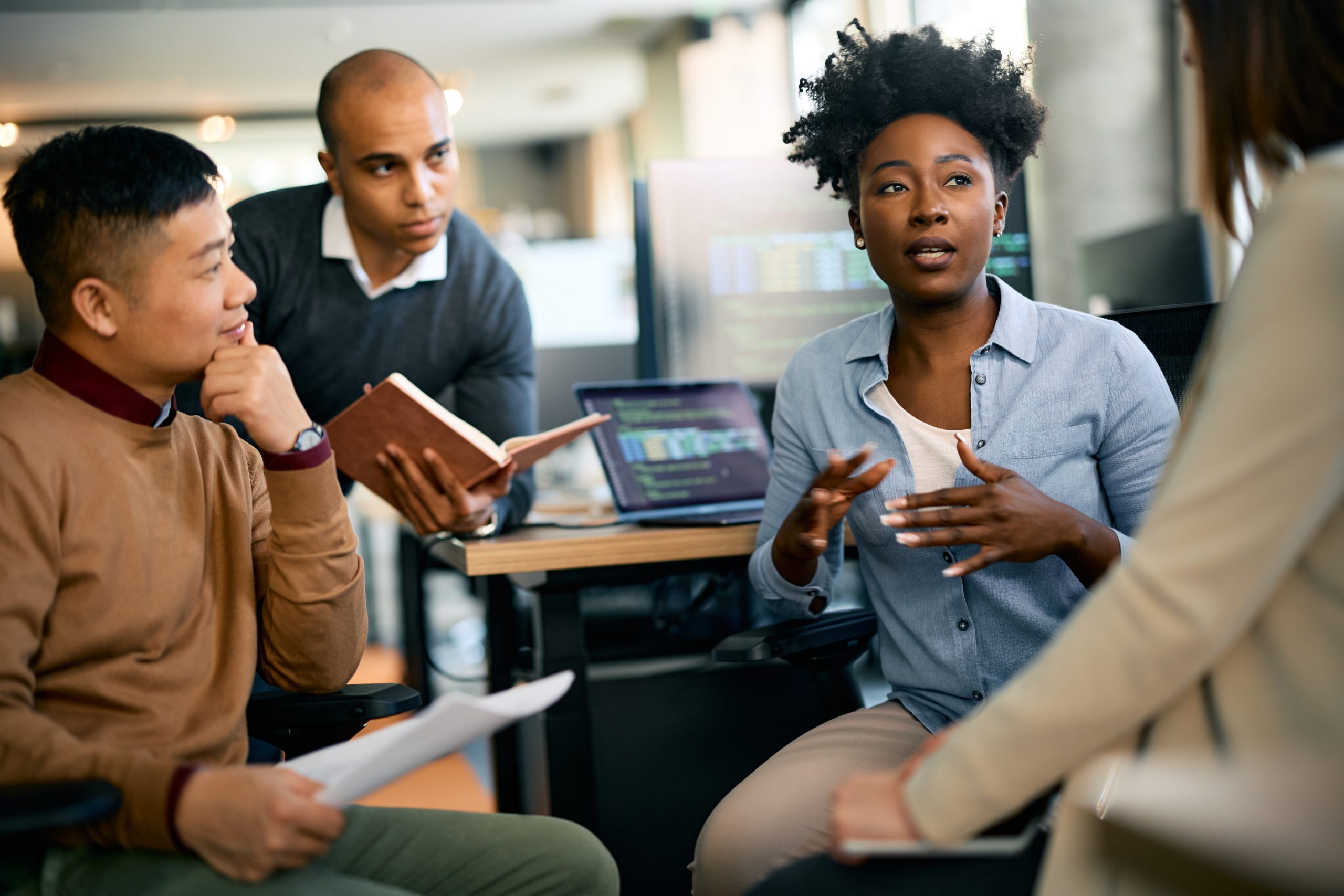 Three people in an office listen to a woman in a blue button-down speaking.