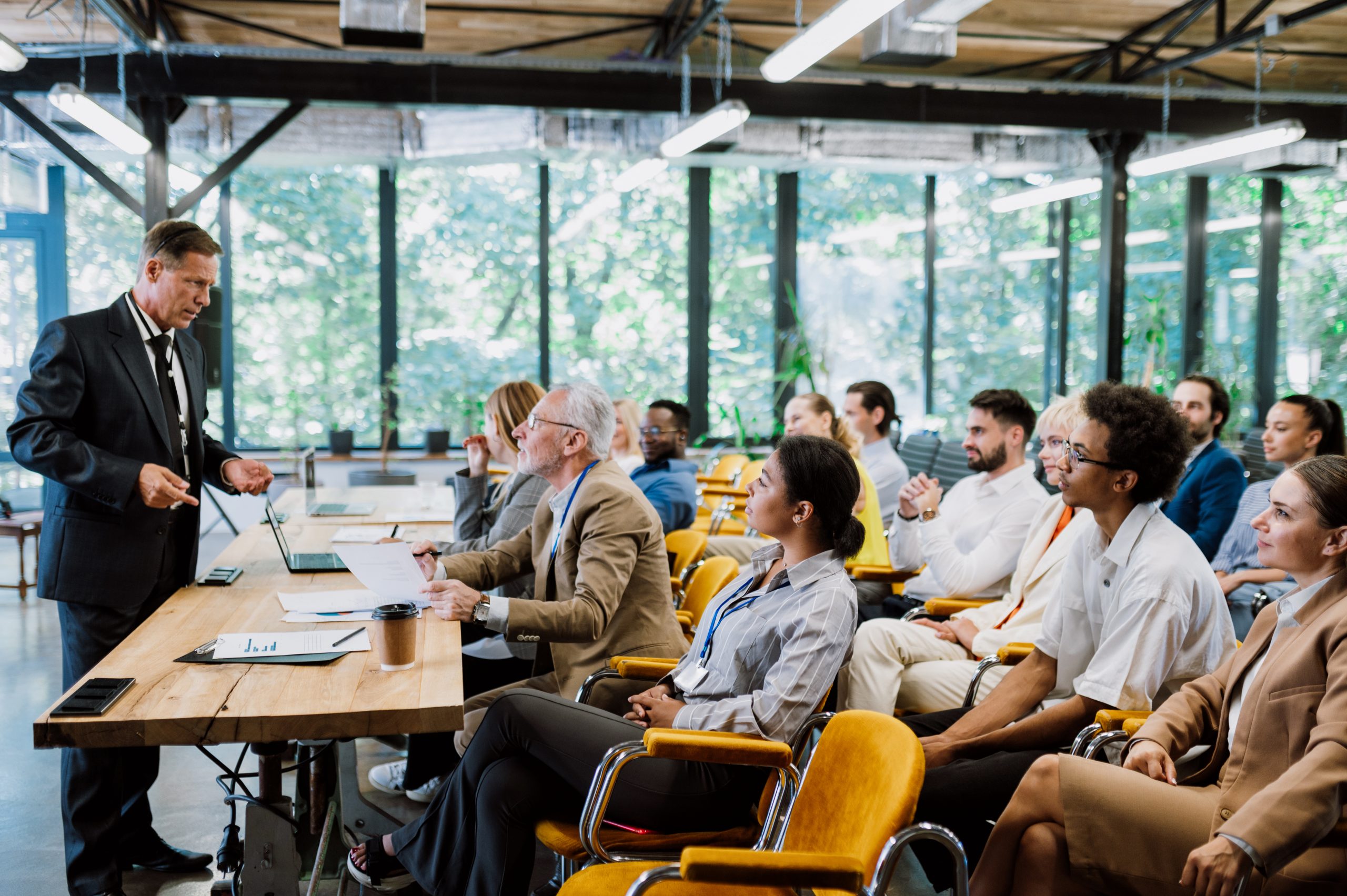 A man in a suit presents to an office of business professionals seated in yellow chairs.