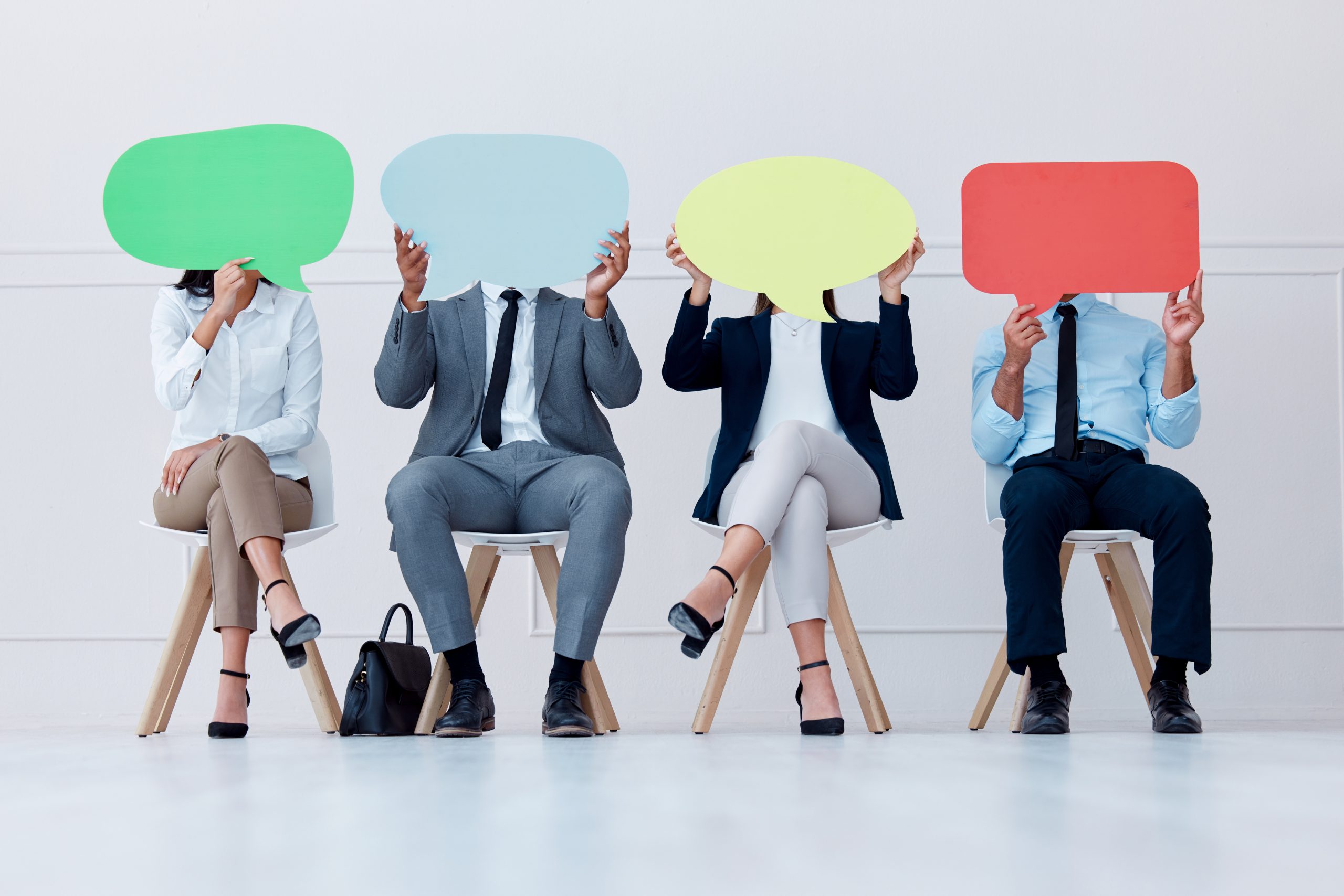Four people in business attire sit in chairs, covering their faces with colorful speech bubble signs.